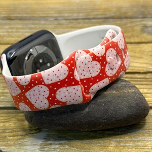 May include: A red and white heart-patterned watch strap with a black watch face. The strap has a repeating pattern of pink hearts and white polka dots. The watch strap is made of a flexible material and is resting on a dark grey stone.