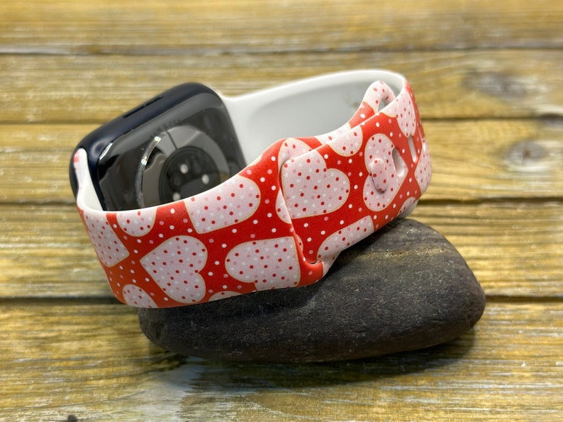 May include: A red and white heart-patterned watch strap with a black watch face. The strap has a repeating pattern of pink hearts and white polka dots. The watch strap is made of a flexible material and is resting on a dark grey stone.