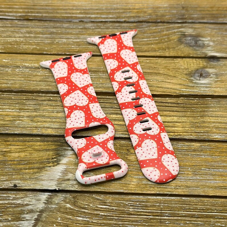 May include: Two red and white patterned watch straps. The straps feature a repeating pattern of white hearts with small white polka dots on a red background. The straps are made of a flexible material and are designed to fit a smartwatch.