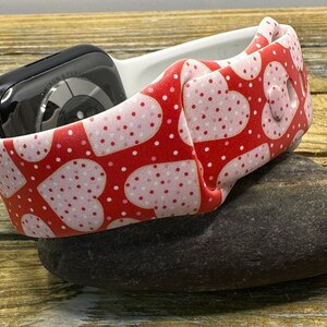 May include: An Apple Watch strap with a red heart pattern and white polka dots. The strap is wrapped around a black Apple Watch. The band is resting on a dark grey stone.