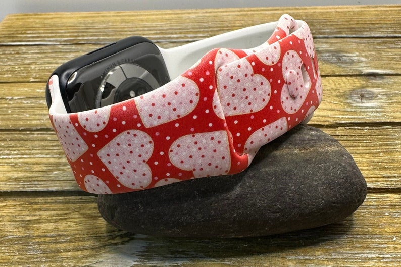 May include: An Apple Watch strap with a red heart pattern and white polka dots. The strap is wrapped around a black Apple Watch. The band is resting on a dark grey stone.