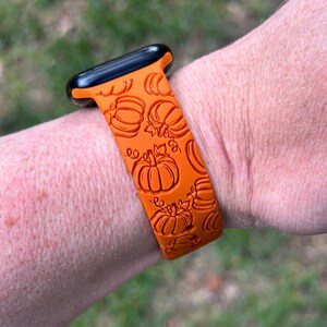 May include: Orange watch band with a pumpkin pattern. The band is made of a flexible material and features a repeating design of carved pumpkins. The watch band is attached to a black watch face.