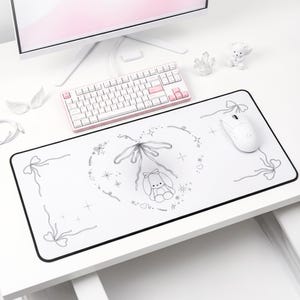 May include: A white desk setup featuring a computer monitor, keyboard, and mouse. A large white mousepad with a black border is decorated with a bunny and bow design. The keyboard has white and pink keys.