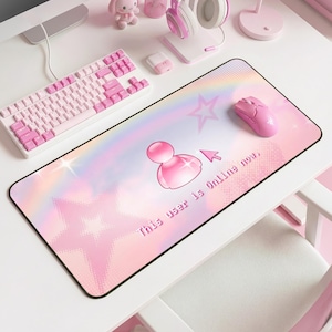 May include: A pink and white computer setup featuring a keyboard, mouse, and headset. A large rectangular mousepad with a pink and rainbow design, a user icon, and the text "This user is Online now." is the focal point.