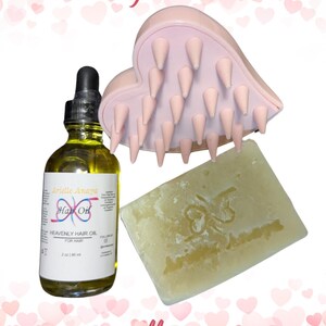May include: A pink heart-shaped scalp massager, a clear glass bottle of hair oil labeled "Arielle Anaya Hair Oil Heavenly Hair Oil for Hair 2 oz | 60 ml", and a white bar of soap with the text "Arielle Anaya" on it. The items are arranged on a white background with pink hearts.