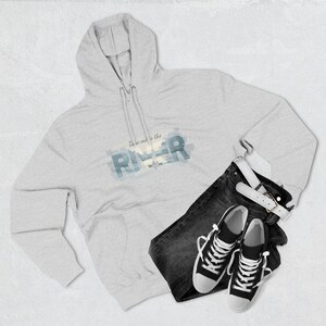 May include: A light gray hoodie with the text "Take me to the River" in blue and white. The hoodie is paired with dark wash jeans, a white belt, and black and white sneakers.