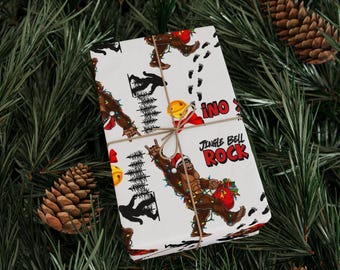 May include: White wrapping paper with a repeating pattern of a Bigfoot character wearing a Santa hat and holding a gift bag. The words "Jingle Bell Rock" are printed on the paper. The paper is tied with twine and surrounded by pine needles and pine cones.