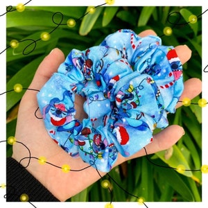 Puede incluir: Scrunchie azul y blanco con un estampado navideño con un personaje de dibujos animados con un gorro de Papá Noel. El estampado también incluye bastones de caramelo y luces navideñas.