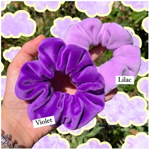 May include: Two velvet scrunchies in shades of purple. The scrunchie on the left is a solid dark purple labeled "Violet". The scrunchie on the right is a light purple ombre scrunchie labeled "Lilac".