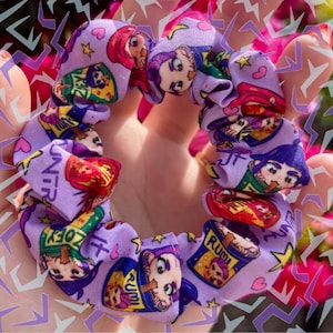 Puede incluir: Un coletero de tela de color morado claro con un patrón repetitivo de personajes de dibujos animados en cajas coloridas con los nombres Zoey y Rumi impresos, intercalados con pequeñas estrellas amarillas y corazones rosas en el material.