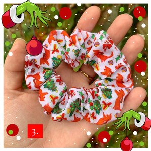Puede incluir: Una goma para el pelo blanca con un estampado navide&ntilde;o con perros rojos, &aacute;rboles de Navidad y adornos. La goma para el pelo se sostiene en una mano, mostrando su dise&ntilde;o. El n&uacute;mero "3." es visible en la esquina inferior izquierda.