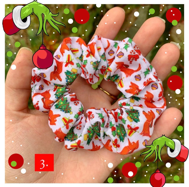 Puede incluir: Una goma para el pelo blanca con un estampado navide&ntilde;o con perros rojos, &aacute;rboles de Navidad y adornos. La goma para el pelo se sostiene en una mano, mostrando su dise&ntilde;o. El n&uacute;mero "3." es visible en la esquina inferior izquierda.