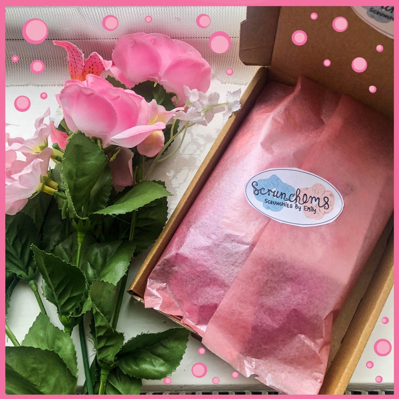 May include: A brown cardboard box with a pink tissue paper lining. The box is open and the tissue paper is folded over. A white oval sticker with the text "Scrunchoms Scrunchies by Emily" is on the tissue paper. The box is surrounded by pink flowers.