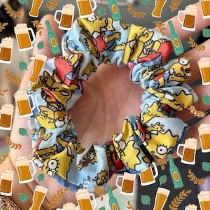May include: A blue and yellow scrunchie featuring a cartoon print of characters from The Simpsons. The scrunchie is surrounded by a pattern of beer mugs and hops.