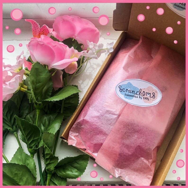 K&ouml;nnte beinhalten: Eine Geschenkbox mit rosa Seidenpapier und einem Aufkleber mit der Aufschrift "Scrunchems Scrunchies By Emily". Rosa Rosen und Gr&uuml;nzeug umgeben die Box, was auf ein Geschenk oder eine Produktpr&auml;sentation hindeutet.