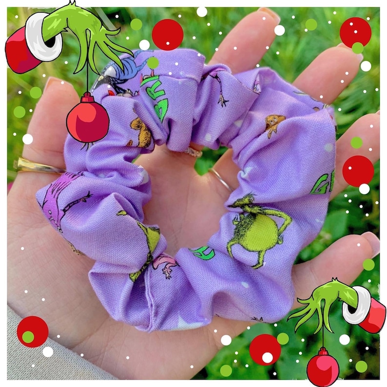 Puede incluir: Una scrunchie lavanda con un patr&oacute;n repetido de un personaje verde y el texto "Grinch". El accesorio para el cabello est&aacute; hecho de tela. Esta scrunchie est&aacute; dise&ntilde;ada para peinar el cabello y es un accesorio festivo.