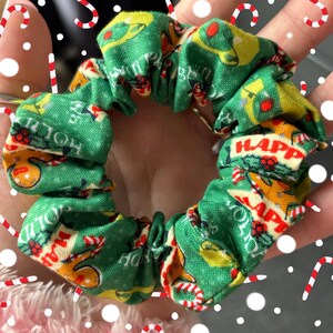 Puede incluir: Scrunchie de Navidad verde y amarillo con las palabras "Felices Fiestas" y "Feliz" repetidas en la tela.