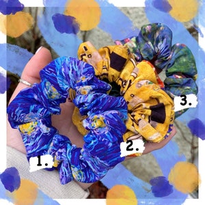 K&ouml;nnte beinhalten: Drei Stoff-Haargummis mit k&uuml;nstlerischen Mustern. Eines ist blau mit einem Sternennacht-Design, ein anderes ist gelb mit einem Geb&auml;udedesign und das dritte ist gr&uuml;n mit einem floralen Design. Die Haargummis werden in einer Hand gehalten.