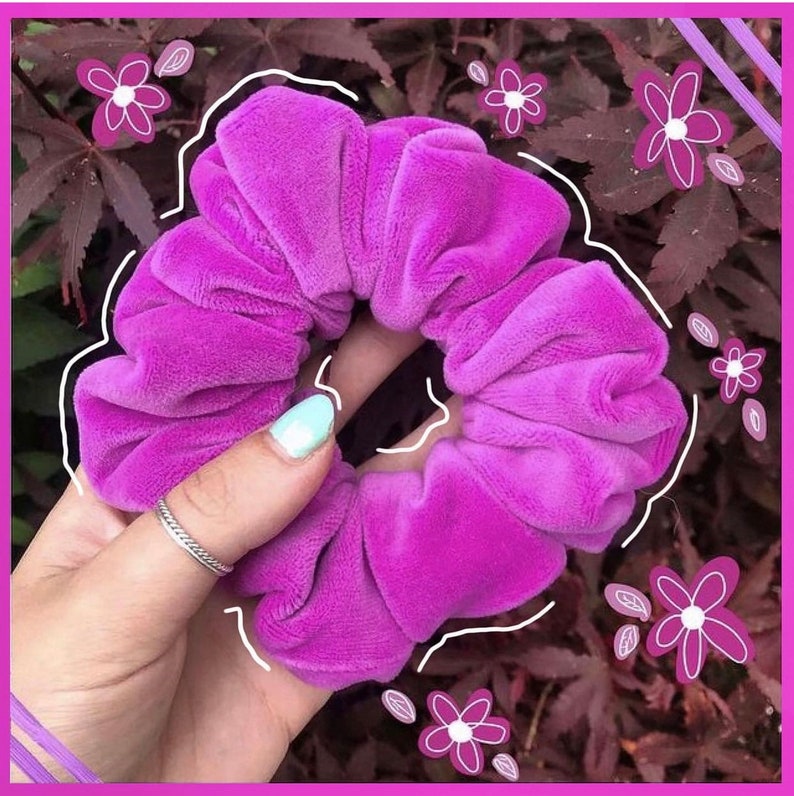 May include: A bright purple velvet scrunchie, a popular hair accessory, is held in a hand against a backdrop of green leaves.