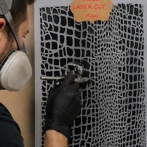 Peut inclure: Un pochoir rectangulaire présentant un motif répétitif de peau de crocodile noire sur un fond gris clair. Une étiquette beige en haut indique Durable LASER CUT Mylar. Le pochoir est utilisé avec un aérographe argenté pour appliquer la peinture.