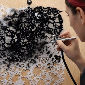 May include: A silver airbrush tool applying black ink over a white, organic, lace-like stencil pattern on a light wood surface. The stencil features intricate, irregular, interconnected shapes that create a high-contrast, textured visual effect.