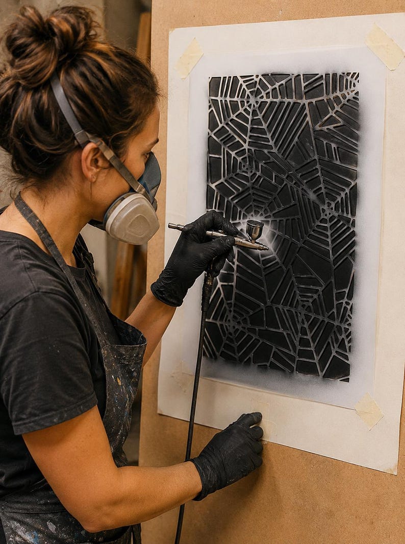 May include: A rectangular stencil featuring a complex, repeating spider web pattern is being used to apply black paint onto a surface with an airbrush tool. The design consists of interconnected geometric web segments, creating a high-contrast visual effect.