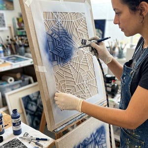 May include: A rectangular canvas mounted on a wooden easel features a white stencil with a geometric spider web pattern. A hand-held airbrush tool applies blue paint over the stencil, creating a gradient effect on the textured canvas surface.
