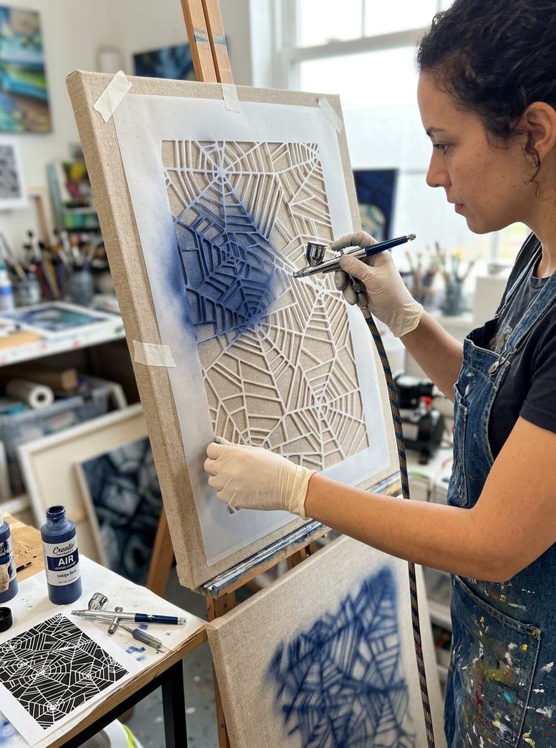 May include: A rectangular canvas mounted on a wooden easel features a white stencil with a geometric spider web pattern. A hand-held airbrush tool applies blue paint over the stencil, creating a gradient effect on the textured canvas surface.