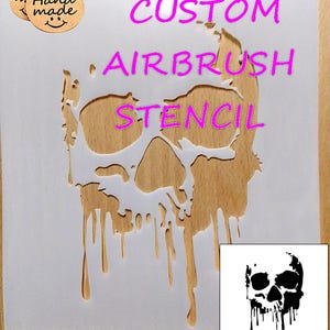 May include: A white airbrush stencil with a skull design and the words "CUSTOM AIRBRUSH STENCIL" in pink. The stencil is on a wooden surface. A small tag says "Hand made".