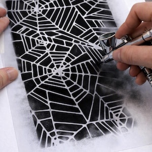 May include: A rectangular stencil featuring a repeating spider web pattern being used with a silver airbrush tool to apply black paint onto a white surface, creating a high-contrast graphic design with sharp lines and soft, sprayed edges.