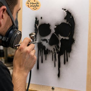 May include: A white rectangular sheet featuring a black spray-painted skull design with dripping paint effects. A circular brown paper tag with the text "Hand made" and a smiley face icon is attached to the top left corner of the white sheet.