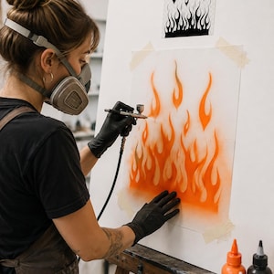 May include: An airbrush tool is used to apply orange paint onto a white surface, creating a flame pattern. The artwork features a stencil of black flames above the painted orange flames. The artist wears a respirator mask and black gloves.