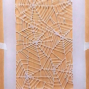 May include: A rectangular stencil featuring a repeating white spider web pattern on a translucent sheet. The design consists of multiple interconnected web centers with radiating lines and concentric segments, creating a detailed geometric network.