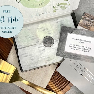 Op de afbeelding: Een grijze kaart met de tekst "Free Gift Note with Every Order" en instructies over hoe je een cadeaukaart aan een bestelling kunt toevoegen. De kaart ligt op een houten oppervlak met een gouden spatel en een doos met een vlinderlogo.
