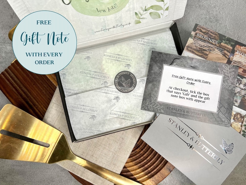 Op de afbeelding: Een grijze kaart met de tekst "Free Gift Note with Every Order" en instructies over hoe je een cadeaukaart aan een bestelling kunt toevoegen. De kaart ligt op een houten oppervlak met een gouden spatel en een doos met een vlinderlogo.