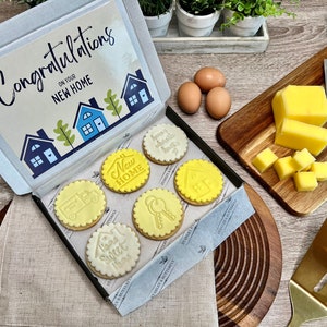 Op de afbeelding: Een cadeaudoos gevuld met zes gedecoreerde koekjes. De koekjes zijn geel en wit met verschillende huis-thema ontwerpen. De doos heeft een kaart bovenop die zegt "Gefeliciteerd met je nieuwe huis".