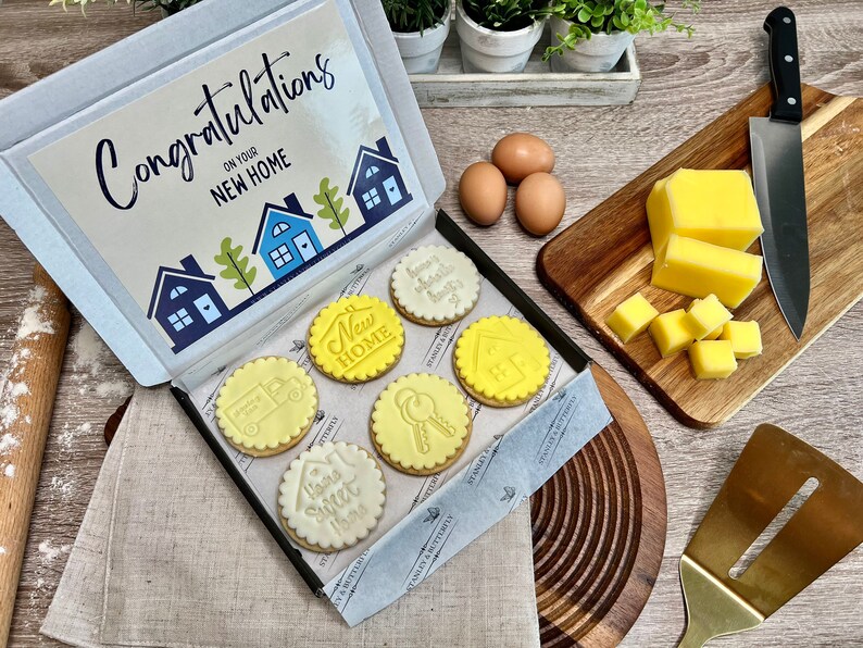 Op de afbeelding: Een cadeaudoos gevuld met zes gedecoreerde koekjes. De koekjes zijn geel en wit met verschillende huis-thema ontwerpen. De doos heeft een kaart bovenop die zegt "Gefeliciteerd met je nieuwe huis".