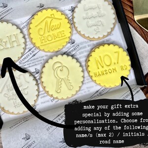 Op de afbeelding: Een set van zes gedecoreerde koekjes met verschillende ontwerpen, waaronder "New Home", "T&H", "Home Sweet Home", een sleutel en "NO.7 HANSOM RD". De koekjes zijn gerangschikt in een doos met een zwarte achtergrond. De tekst "make your gift extra special by adding some personalisation. Choose from adding any of the following... name/s (max 2) / initials / road name" staat op een zwarte achtergrond.