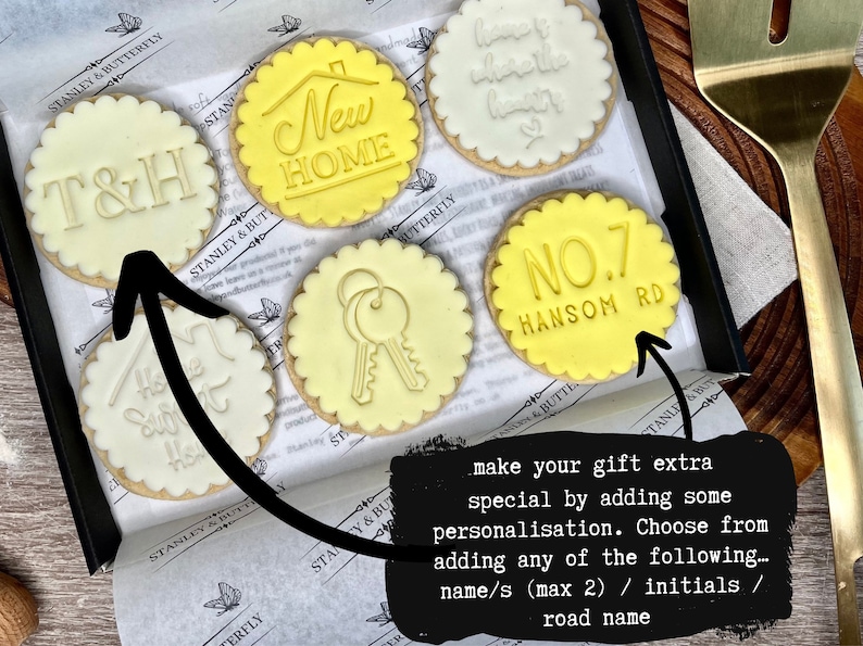 Op de afbeelding: Een set van zes gedecoreerde koekjes met verschillende ontwerpen, waaronder "New Home", "T&H", "Home Sweet Home", een sleutel en "NO.7 HANSOM RD". De koekjes zijn gerangschikt in een doos met een zwarte achtergrond. De tekst "make your gift extra special by adding some personalisation. Choose from adding any of the following... name/s (max 2) / initials / road name" staat op een zwarte achtergrond.