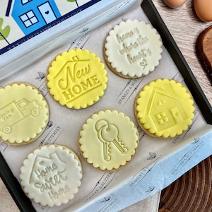 Op de afbeelding: Een doos met zes gedecoreerde koekjes met een nieuw-thuis-thema. De koekjes zijn rond en hebben gegolfde randen. De ontwerpen omvatten een huis, sleutels, een verhuiswagen en de zinnen "Home Sweet Home", "New Home" en "Home is where the heart is".
