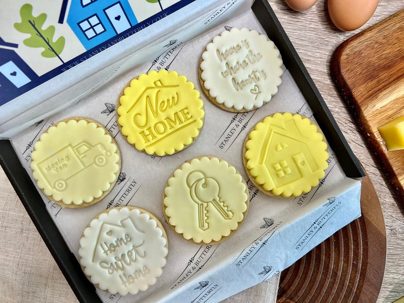 Op de afbeelding: Een doos met zes gedecoreerde koekjes met een nieuw-thuis-thema. De koekjes zijn rond en hebben gegolfde randen. De ontwerpen omvatten een huis, sleutels, een verhuiswagen en de zinnen "Home Sweet Home", "New Home" en "Home is where the heart is".