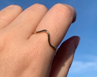 May include: A gold-colored wavy ring worn on a finger against a blue sky background.