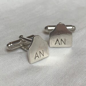May include: A pair of silver cufflinks with a unique design. Each cufflink features a silver tag with the initials "AN" engraved on it.