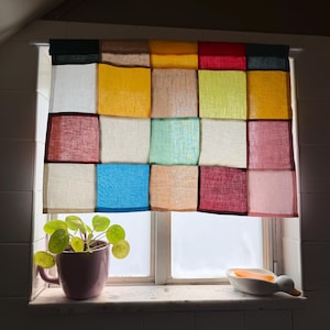 Op de afbeelding: Een raamgordijn met een patchworkontwerp van vierkante stoffen panelen in diverse kleuren, waaronder geel, blauw, rood, groen, wit en beige. Het gordijn hangt aan een witte roede voor een raam, met een groene plant in een roze mok ernaast.