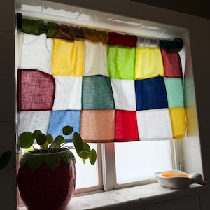 May include: A window valance featuring a patchwork design of square fabric panels in various colors including red, yellow, green, blue, white, and brown. The curtain is gathered on a black rod and hangs over a window pane with a light texture.