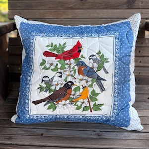 May include: Square cushion with a detailed print of several birds, including a red cardinal, amidst white flowers and green leaves. The cushion has a white centre with a blue border and a blue polka dot pattern on the edges.