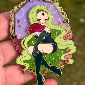 May include: Enamel pin featuring a seated anime-style character with long green hair, a black outfit, and a red fan. The pin has a gold frame with a purple background and floral accents. The character has a crescent moon on their forehead.