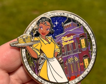 May include: A gold-toned enamel pin featuring Tiana from Disney's The Princess and the Frog. Tiana is wearing a yellow dress and white apron and holding a plate of pancakes. The background features a purple building with the text "Tiana's Palace".