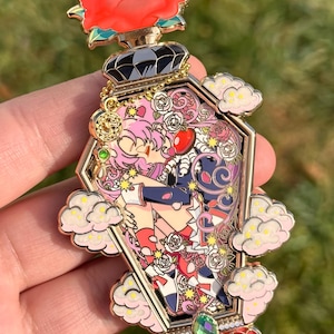 May include: Gold-tone enamel pin depicting a pink-haired character in a coffin with a red rose on top. The coffin is surrounded by white clouds and roses. The character is wearing a red and white dress and a blue jacket.