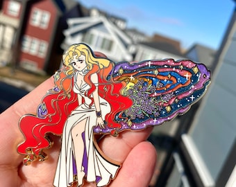 May include: A gold-tone enamel pin featuring a woman with long red hair and a white dress. Her hair is flowing behind her, forming a galaxy with stars and purple flowers.
